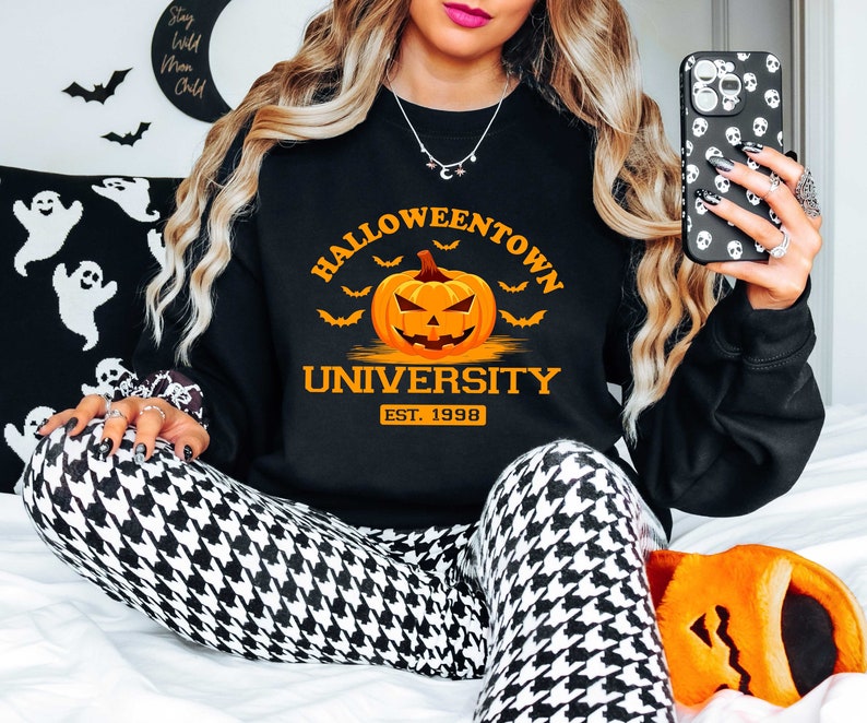 May include: Black sweatshirt with the text "Halloweentown University Est. 1998" and a cartoon orange pumpkin with a scary face. The sweatshirt is worn by a person with long blonde hair and is sitting on a white surface.