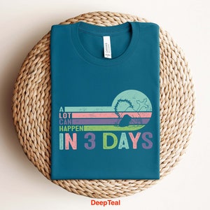 Puede incluir: Una camiseta azul turquesa con un gr&aacute;fico de estilo retro que dice "Mucho puede pasar en 3 d&iacute;as". El gr&aacute;fico presenta un dise&ntilde;o a rayas de colores con una silueta de una persona buceando en el agua.