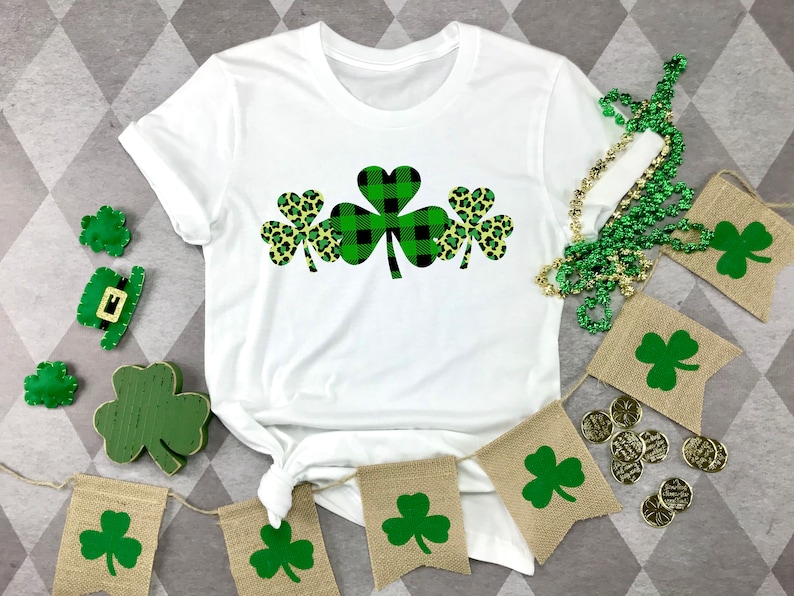May include: White t-shirt with a green, black, and yellow shamrock design. The shamrock is made up of three individual shamrocks, one with a black and white plaid pattern, and two with a leopard print pattern.