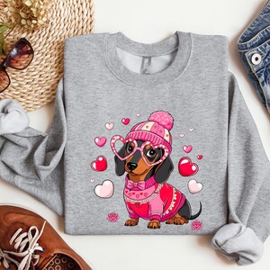 May include: A gray sweatshirt with a cute cartoon dachshund wearing a pink hat and heart-shaped glasses. The dachshund is surrounded by pink hearts and is holding a pink and red scarf. The sweatshirt is perfect for Valentine's Day.