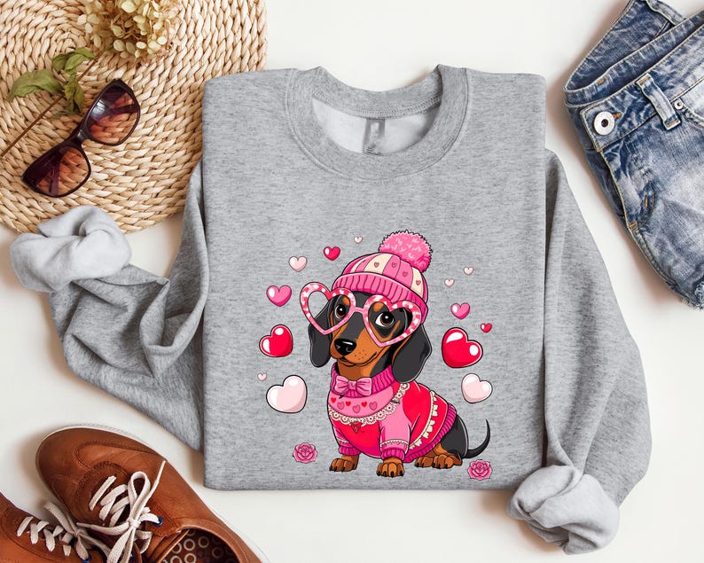 May include: A gray sweatshirt with a cute cartoon dachshund wearing a pink hat and heart-shaped glasses. The dachshund is surrounded by pink hearts and is holding a pink and red scarf. The sweatshirt is perfect for Valentine's Day.