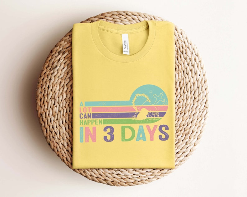 Puede incluir: Una camiseta amarilla con un gr&aacute;fico de estilo retro que dice "Muchas cosas pueden pasar en 3 d&iacute;as". El gr&aacute;fico presenta un fondo a rayas de colores y una silueta de una persona con una corona de espinas.