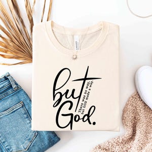 May include: Cream-colored t-shirt with the text "but God" in a black script font, accompanied by a cross graphic. The phrase "There was no way, but GOD made a way" is also printed on the shirt. The shirt is folded and displayed with jeans and white shoes.