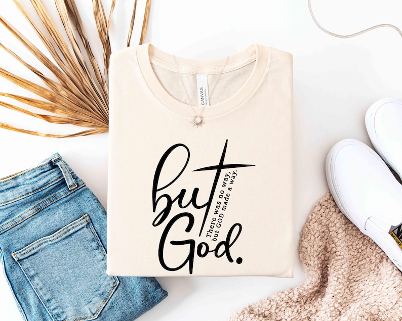 May include: Cream-colored t-shirt with the text "but God" in a black script font, accompanied by a cross graphic. The phrase "There was no way, but GOD made a way" is also printed on the shirt. The shirt is folded and displayed with jeans and white shoes.