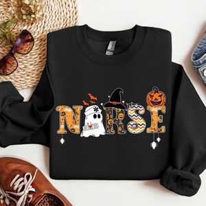 May include: Black sweatshirt with a Halloween-themed "NURSE" graphic. The graphic features a ghost, a witch's hat, a pumpkin, and other Halloween elements.