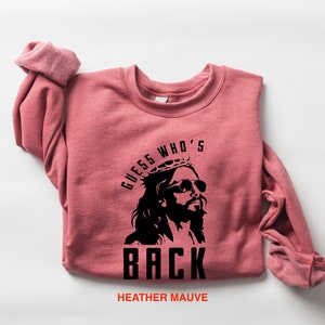 Puede incluir: Una sudadera de color malva con un gr&aacute;fico negro de Jes&uacute;s con gafas de sol y una corona de espinas. El texto "GUESS WHO'S BACK" est&aacute; impreso debajo de la imagen.