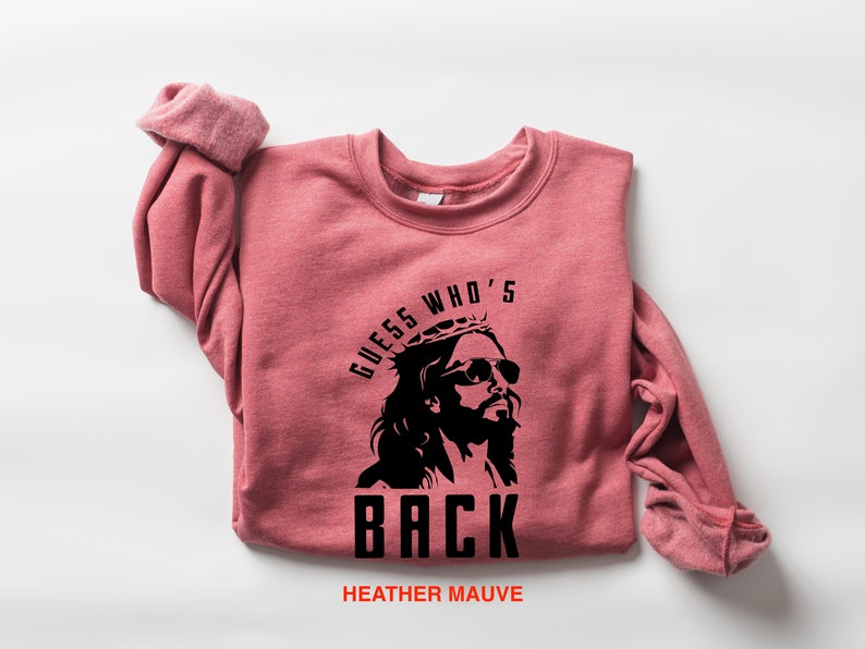 Puede incluir: Una sudadera de color malva con un gr&aacute;fico negro de Jes&uacute;s con gafas de sol y una corona de espinas. El texto "GUESS WHO'S BACK" est&aacute; impreso debajo de la imagen.