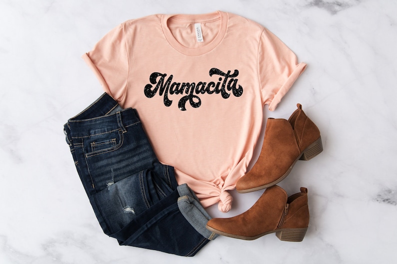 Pu&ograve; includere: Una maglietta color pesca con la scritta "Mamacita" in nero, con un lettering in stile retr&ograve;. La maglietta &egrave; abbinata a jeans in denim scuro e stivaletti marroni. Gli articoli sono disposti su una superficie di marmo bianco.