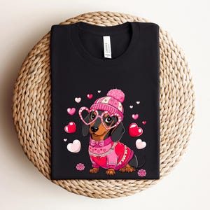 May include: Black t-shirt with a cartoon dachshund wearing a pink hat, pink heart-shaped glasses, and a pink sweater with hearts. The dachshund is surrounded by pink hearts.