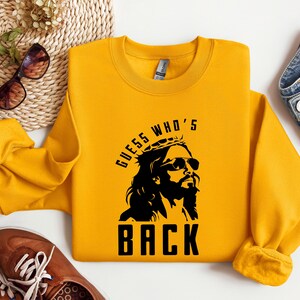 Puede incluir: Sudadera amarilla con un gr&aacute;fico negro de Jes&uacute;s con gafas de sol y una corona de espinas. El texto "Guess Who's Back" est&aacute; impreso debajo del gr&aacute;fico.
