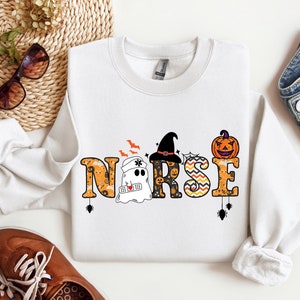 May include: White sweatshirt with a Halloween-themed design featuring a ghost, a witch, and a pumpkin. The design spells out the word "NURSE" with Halloween-themed embellishments.