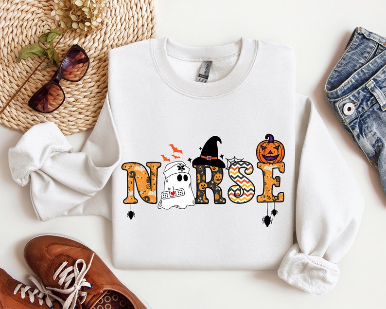 May include: White sweatshirt with a Halloween-themed design featuring a ghost, a witch, and a pumpkin. The design spells out the word "NURSE" with Halloween-themed embellishments.