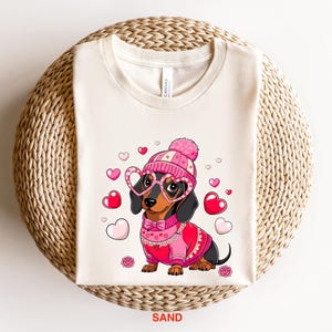 May include: A white t-shirt with a cartoon image of a dachshund wearing a pink and white hat, pink heart-shaped glasses, and a pink sweater with a white lace trim. The dachshund is surrounded by pink hearts.