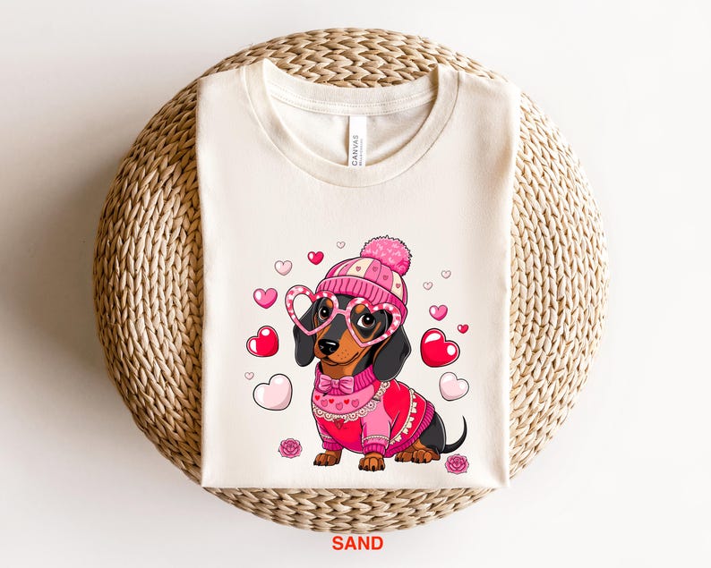 May include: A white t-shirt with a cartoon image of a dachshund wearing a pink and white hat, pink heart-shaped glasses, and a pink sweater with a white lace trim. The dachshund is surrounded by pink hearts.