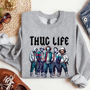 Peut inclure: Un sweat-shirt gris chin&eacute; avec l'inscription "THUG LIFE" en noir, dans une police gothique. Le sweat-shirt pr&eacute;sente un graphisme de cinq personnages de dessins anim&eacute;s portant des sweats &agrave; capuche et des vestes. Les personnages sont dans des tons de bleu, de rouge, de vert et de blanc.