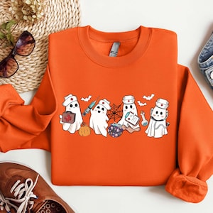 May include: Orange sweatshirt with a graphic of five cartoon ghosts dressed as nurses. The ghosts are holding medical supplies like a syringe, a first aid kit, and a clipboard. The ghosts are wearing red crosses on their hats.