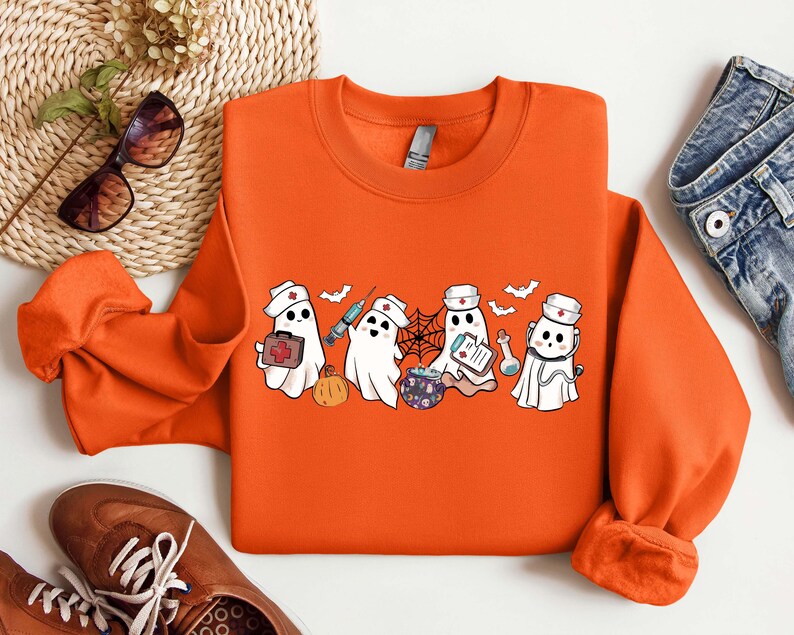 May include: Orange sweatshirt with a graphic of five cartoon ghosts dressed as nurses. The ghosts are holding medical supplies like a syringe, a first aid kit, and a clipboard. The ghosts are wearing red crosses on their hats.