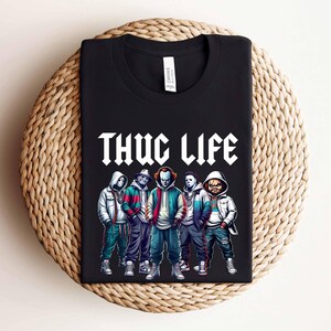 Peut inclure: T-shirt noir avec l'inscription blanche "THUG LIFE", orn&eacute; d'un graphisme de cinq personnages en sweats &agrave; capuche et vestes. Les personnages sont de style cartoon, sur le th&egrave;me de l'horreur.