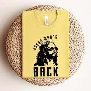 Puede incluir: Camiseta amarilla con un gr&aacute;fico negro de una persona con una corona de espinas y gafas de sol. El texto "Guess Who's Back" est&aacute; impreso debajo del gr&aacute;fico.