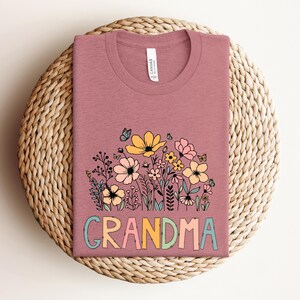 Puede incluir: Una camiseta malva con un dise&ntilde;o floral y la palabra "GRANDMA" en letras de colores.