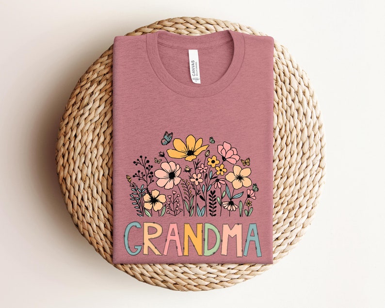 Puede incluir: Una camiseta malva con un dise&ntilde;o floral y la palabra "GRANDMA" en letras de colores.