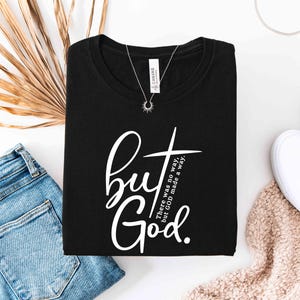 May include: Black t-shirt with the text "but God" in a white script font and a cross graphic. The shirt is folded and displayed with blue jeans, white shoes, and a necklace.