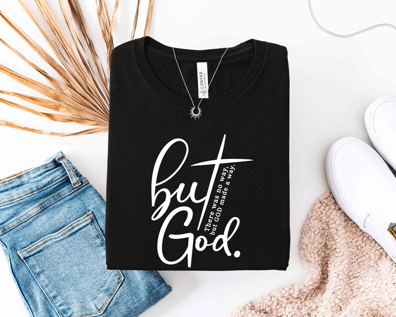 May include: Black t-shirt with the text "but God" in a white script font and a cross graphic. The shirt is folded and displayed with blue jeans, white shoes, and a necklace.