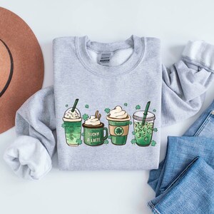 May include: A gray sweatshirt with a graphic of four green drinks with shamrock designs and the text "Lucky a Latte".