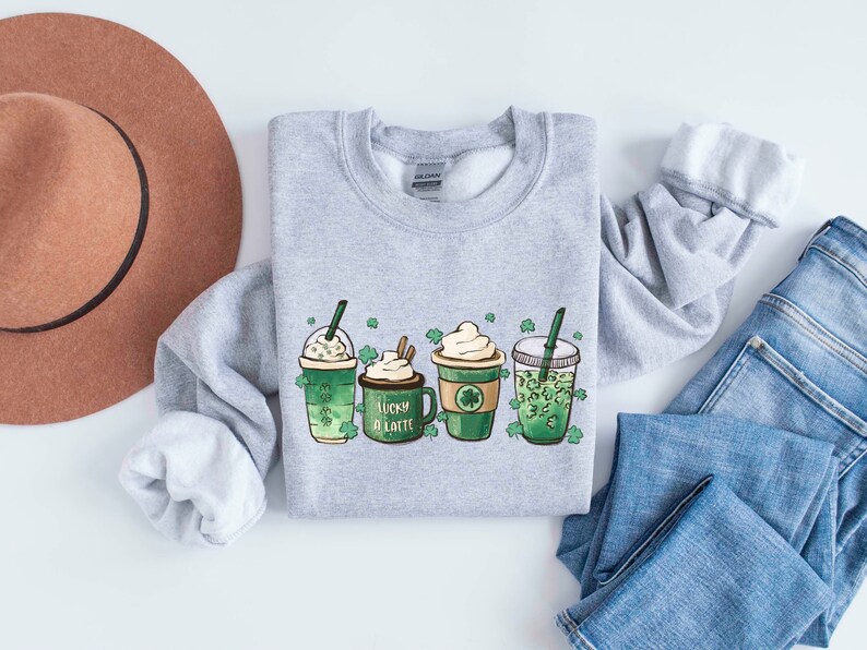 May include: A gray sweatshirt with a graphic of four green drinks with shamrock designs and the text "Lucky a Latte".