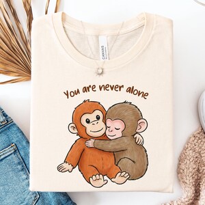 May include: Cream-colored t-shirt featuring a cartoon illustration of two hugging monkeys. The text "You are never alone" is printed above the monkeys in brown. The shirt is folded and displayed on a white surface.