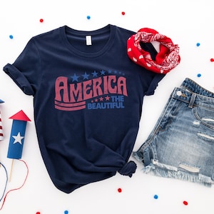Puede incluir: Una camiseta azul marino con un gr&aacute;fico rojo, blanco y azul que dice "America the Beautiful". La camiseta est&aacute; colocada sobre una superficie blanca con confeti rojo, blanco y azul, una bandana roja y unos pantalones cortos de mezclilla.