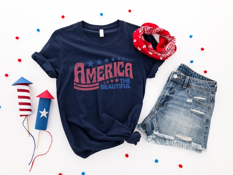Puede incluir: Una camiseta azul marino con un gr&aacute;fico rojo, blanco y azul que dice "America the Beautiful". La camiseta est&aacute; colocada sobre una superficie blanca con confeti rojo, blanco y azul, una bandana roja y unos pantalones cortos de mezclilla.