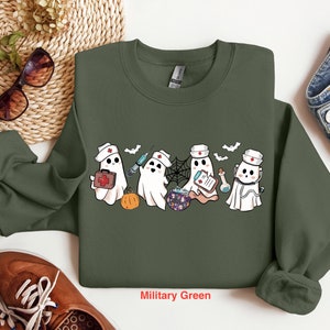 May include: A military green sweatshirt with a graphic of four ghosts dressed as nurses. The ghosts are holding medical supplies and a cauldron. The text "Military Green" is printed below the graphic.