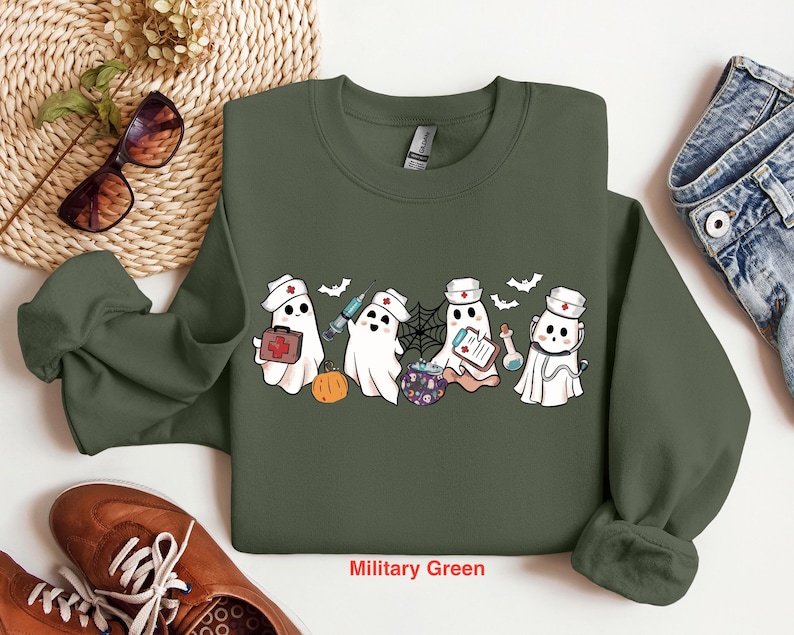 May include: A military green sweatshirt with a graphic of four ghosts dressed as nurses. The ghosts are holding medical supplies and a cauldron. The text "Military Green" is printed below the graphic.