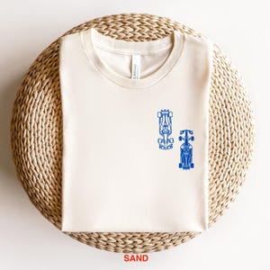 Peut inclure: Un t-shirt blanc avec un motif bleu de deux voitures de course sur le devant. Le t-shirt est sur un tapis de paille tiss&eacute;.