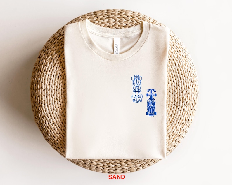 Peut inclure: Un t-shirt blanc avec un motif bleu de deux voitures de course sur le devant. Le t-shirt est sur un tapis de paille tiss&eacute;.