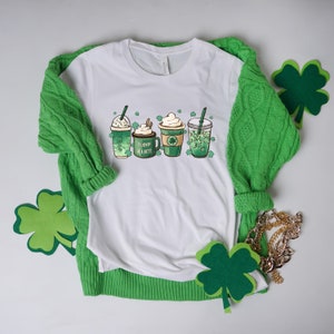 May include: White t-shirt with a graphic of four green coffee drinks with shamrocks and the text "Lucky a Latte". The shirt is displayed on a green sweater with shamrock cutouts.
