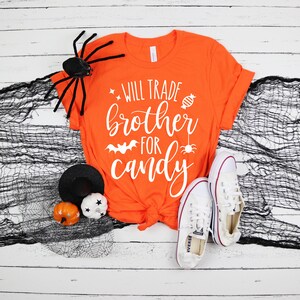 Puede incluir: Camiseta naranja con el texto blanco "Will Trade Brother For Candy". La camiseta se combina con zapatillas blancas, una ara&ntilde;a negra y decoraciones de Halloween. Un dise&ntilde;o divertido y festivo para la temporada de Halloween.