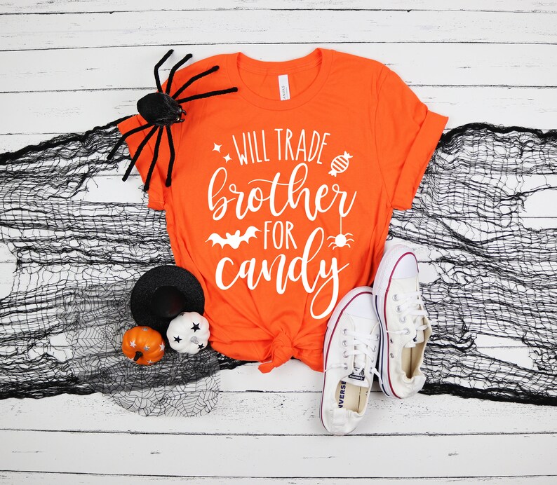 Puede incluir: Camiseta naranja con el texto blanco "Will Trade Brother For Candy". La camiseta se combina con zapatillas blancas, una ara&ntilde;a negra y decoraciones de Halloween. Un dise&ntilde;o divertido y festivo para la temporada de Halloween.