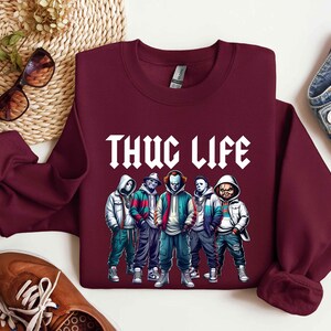Peut inclure: Un sweat-shirt bordeaux avec le texte blanc "THUG LIFE" et un graphisme de cinq personnages de dessins anim&eacute;s en vestes et sweats &agrave; capuche. Les personnages ont un th&egrave;me d'horreur.