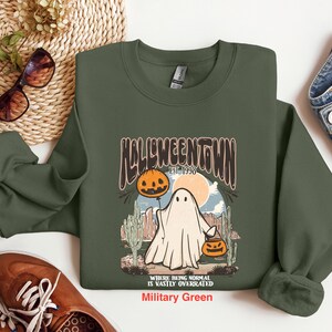 May include: A green sweatshirt with a graphic of a ghost holding a jack-o-lantern. The graphic says "Halloweentown Est. 1998" and "Where being normal is vastly overrated". The sweatshirt is in the color "Military Green".