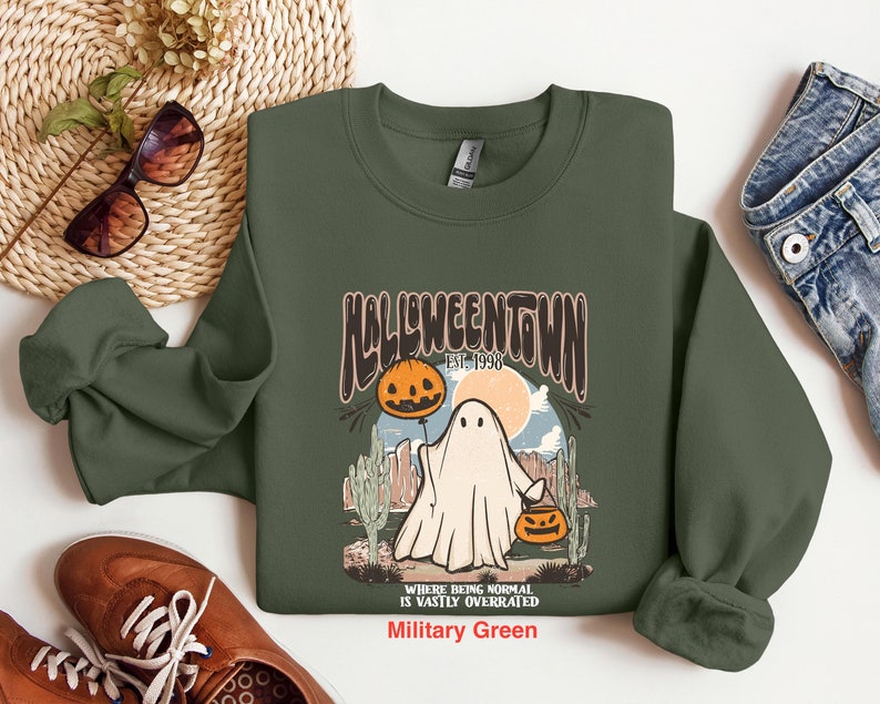 May include: A green sweatshirt with a graphic of a ghost holding a jack-o-lantern. The graphic says "Halloweentown Est. 1998" and "Where being normal is vastly overrated". The sweatshirt is in the color "Military Green".