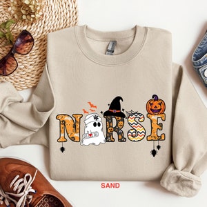 May include: A tan sweatshirt with a Halloween-themed design featuring the word "NURSE" with a ghost, witch hat, and pumpkin. The word "NURSE" is decorated with orange, black, and white patterns.