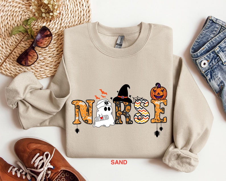 May include: A tan sweatshirt with a Halloween-themed design featuring the word "NURSE" with a ghost, witch hat, and pumpkin. The word "NURSE" is decorated with orange, black, and white patterns.