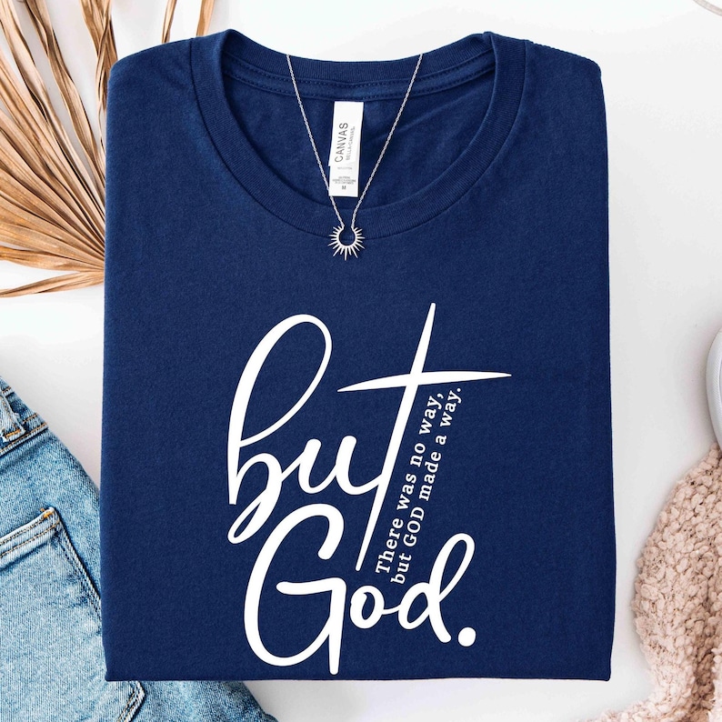 May include: Navy blue t-shirt with the words "but God" in a white script font and a cross graphic. The text "There was no way, but GOD made a way" is also printed on the shirt. A silver necklace with a sunburst pendant is also visible.