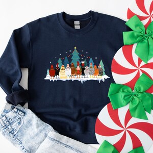 May include: A navy blue sweatshirt with a Christmas-themed design featuring a group of chickens standing in front of a decorated Christmas tree. The chickens are wearing festive hats and scarves.