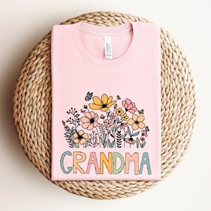Puede incluir: Camiseta rosa con un dise&ntilde;o floral y la palabra "GRANDMA" en letras de colores.
