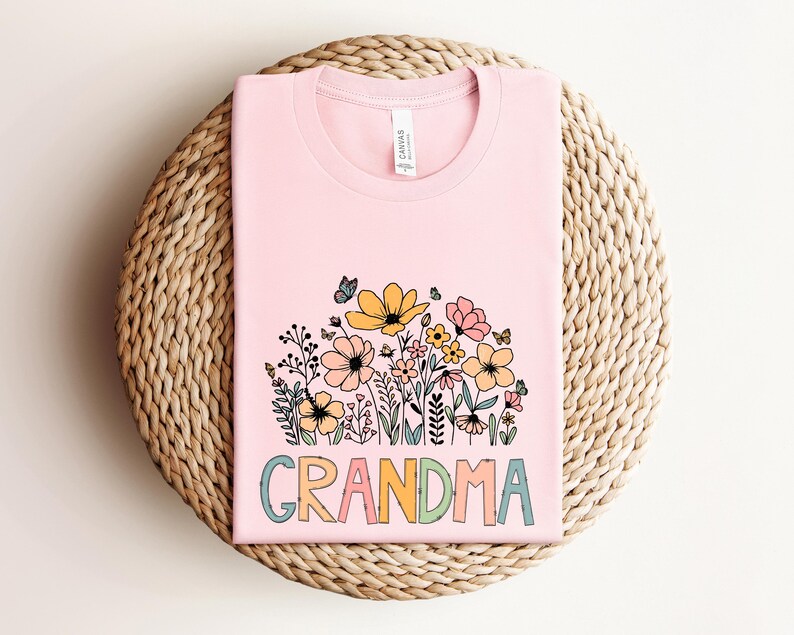 Puede incluir: Camiseta rosa con un dise&ntilde;o floral y la palabra "GRANDMA" en letras de colores.