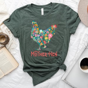 Puede incluir: Camiseta verde oscuro con un dise&ntilde;o floral de gallina. La gallina est&aacute; compuesta por flores coloridas, con el texto "MOTHER HEN" en coral debajo. La camiseta est&aacute; anudada a la cintura.