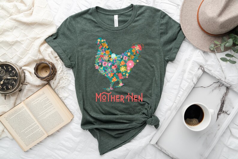 Puede incluir: Camiseta verde oscuro con un dise&ntilde;o floral de gallina. La gallina est&aacute; compuesta por flores coloridas, con el texto "MOTHER HEN" en coral debajo. La camiseta est&aacute; anudada a la cintura.